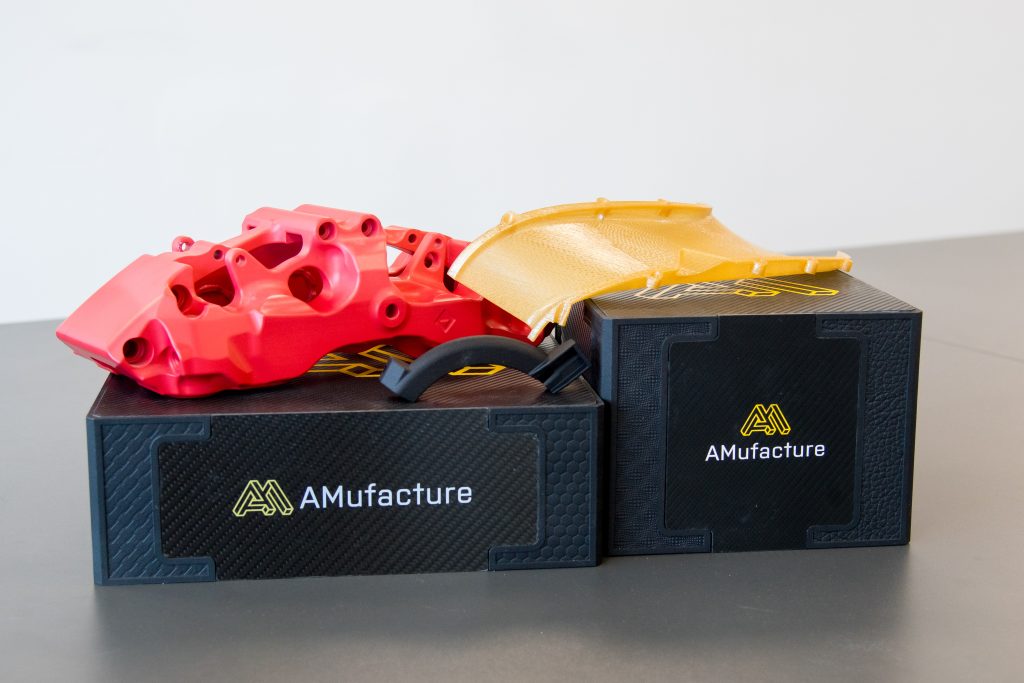 Three 3D printed components on top of two custom made AMufacture boxes. One component is bright red, one is yellow and the third is black.