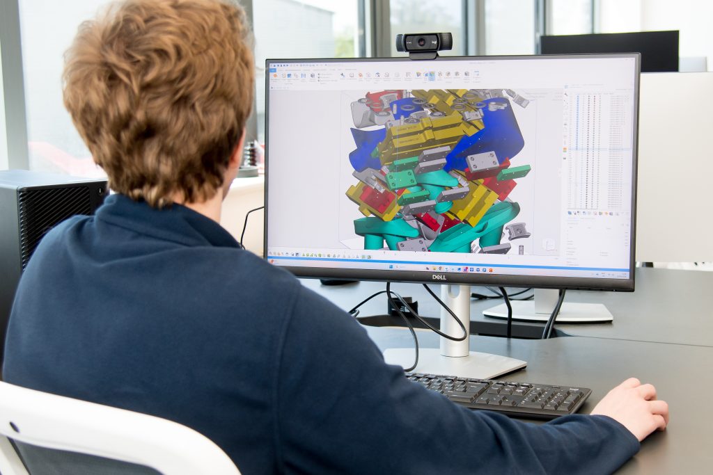 A person sat at a computer monitor. The screen shows a CGI render of various 3D printed components.