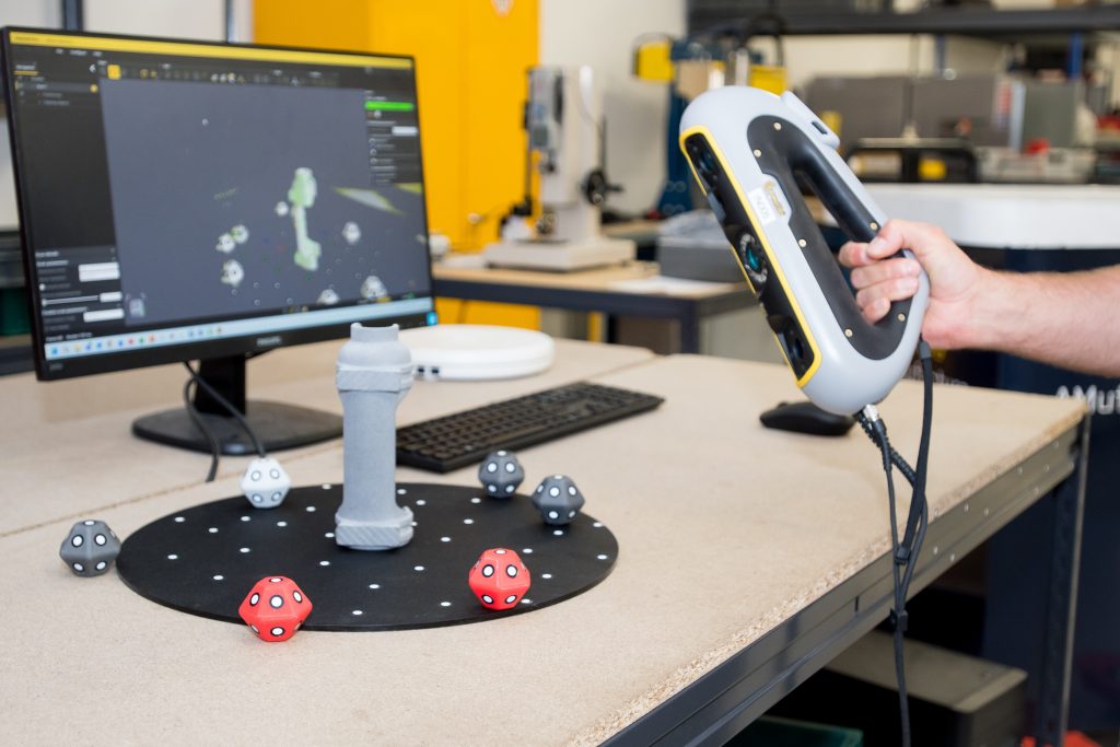 A handheld scanner pointed at a 3D printed component. The computer monitor behind shows the rendered image from the scanner.