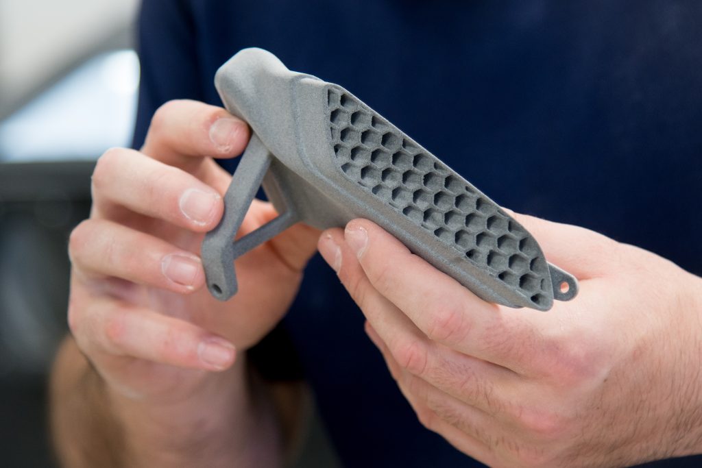 A pair of hands holding a 3D printed component. The component is grey and has a long cavity in the side that is filled with a honeycomb structure.