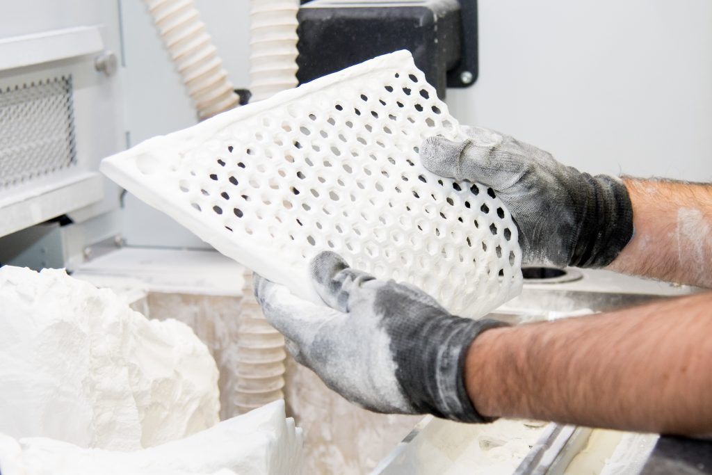 A worker's hands holding a 3D printed component. The component is an off-centre square with a honeycomb structure in its middle. It is covered in a white powder.