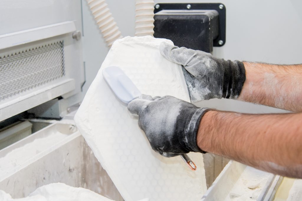 A pair of gloved hands holing up a 3D printed component. The component is covered in a fine white powder and is being brushed over with a small rubber spatula.