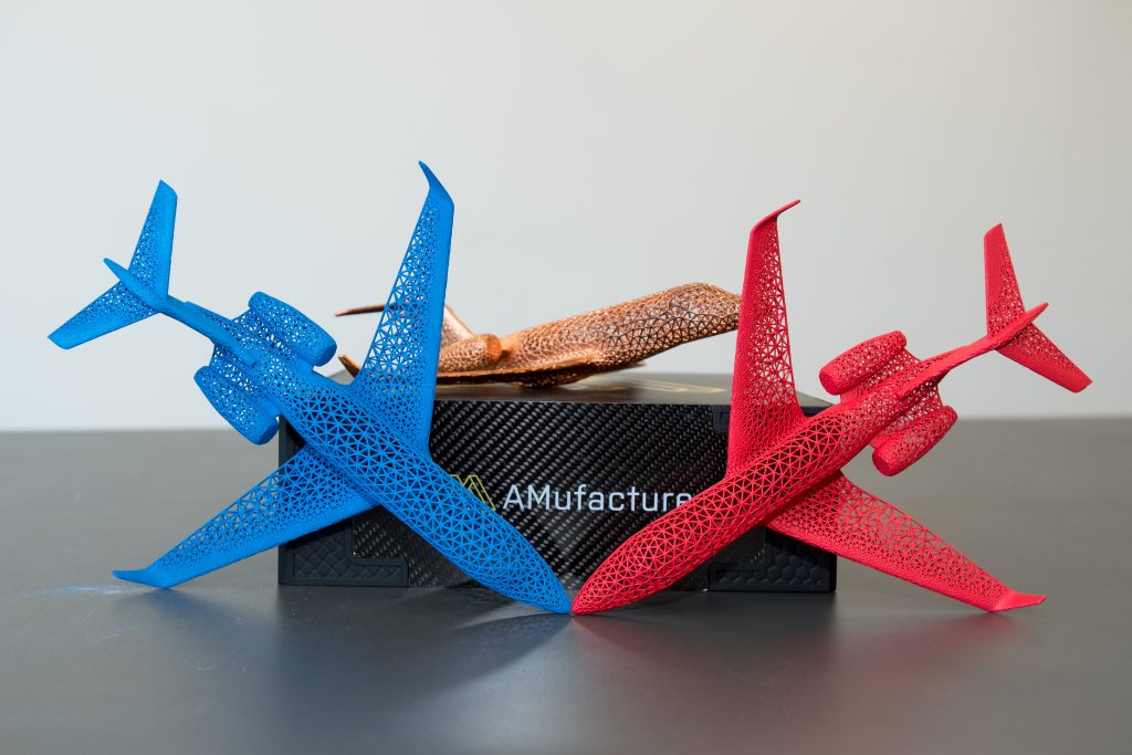 A grey 3D printed lattice manifold with branching arms, displayed on a plinth.