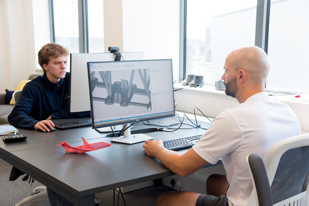 Two people sat facing each other at a desk. Both are working on computers, the screen facing shows a CGI render of a component.