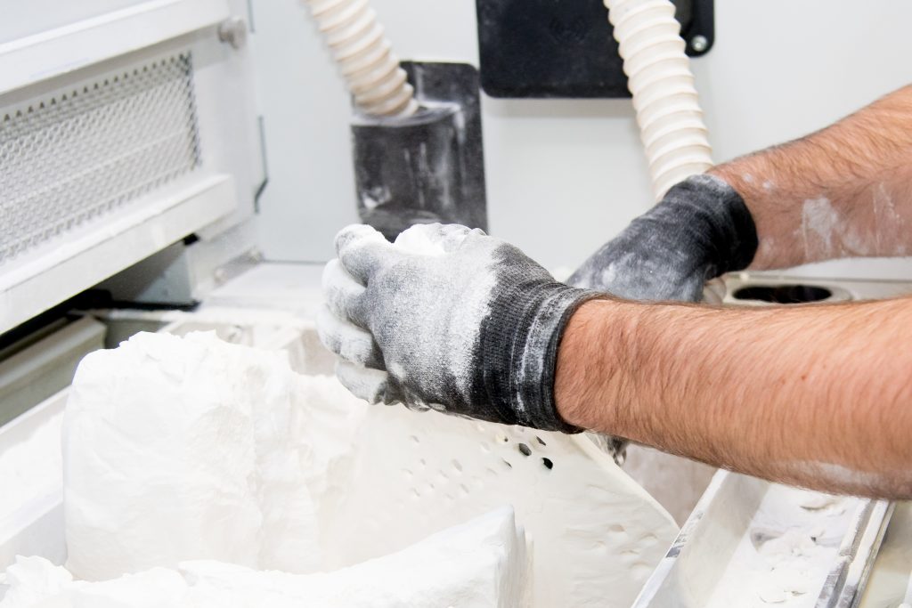 A technician in gloves lifting printed parts out of a bed of loose white nylon powder inside an SLS printer.