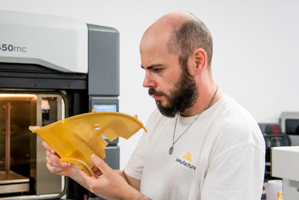 A technician examining a translucent amber printed part in front of an FDM printer.