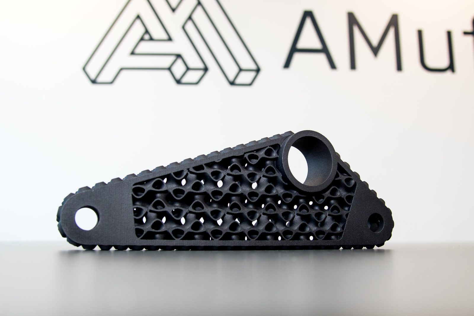 Black 3D printed lightweight arm with an internal lattice structure, in front of the AMufacture logo