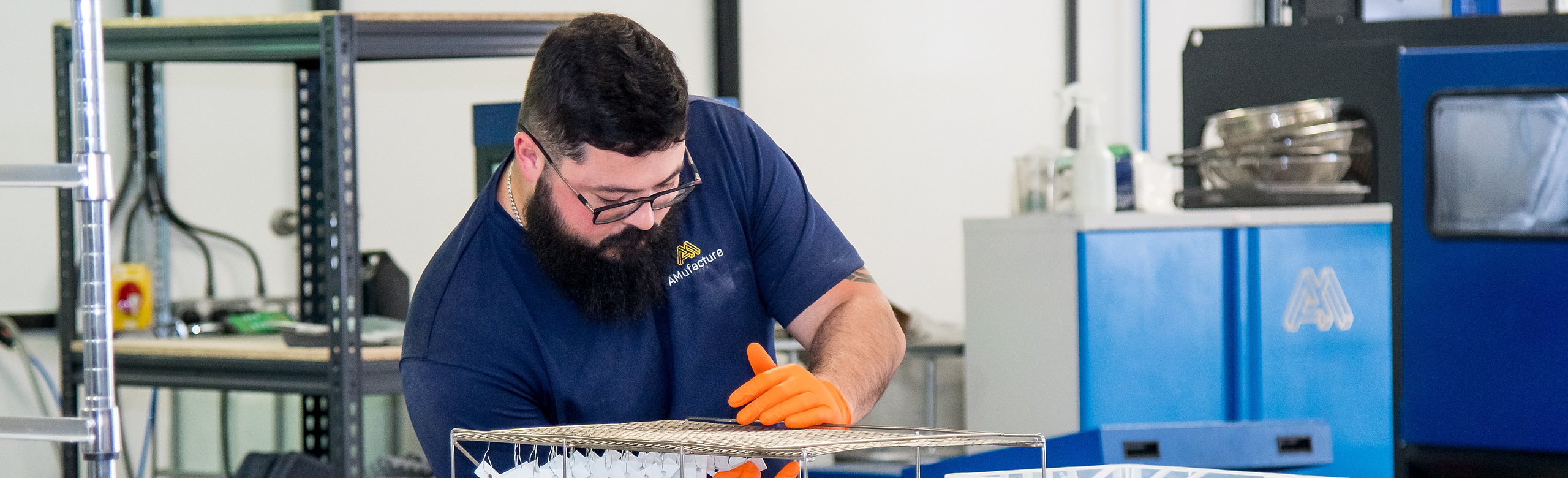 An AMufacture technician in orange gloves handling a rack of finished 3D printed components in the workshop.