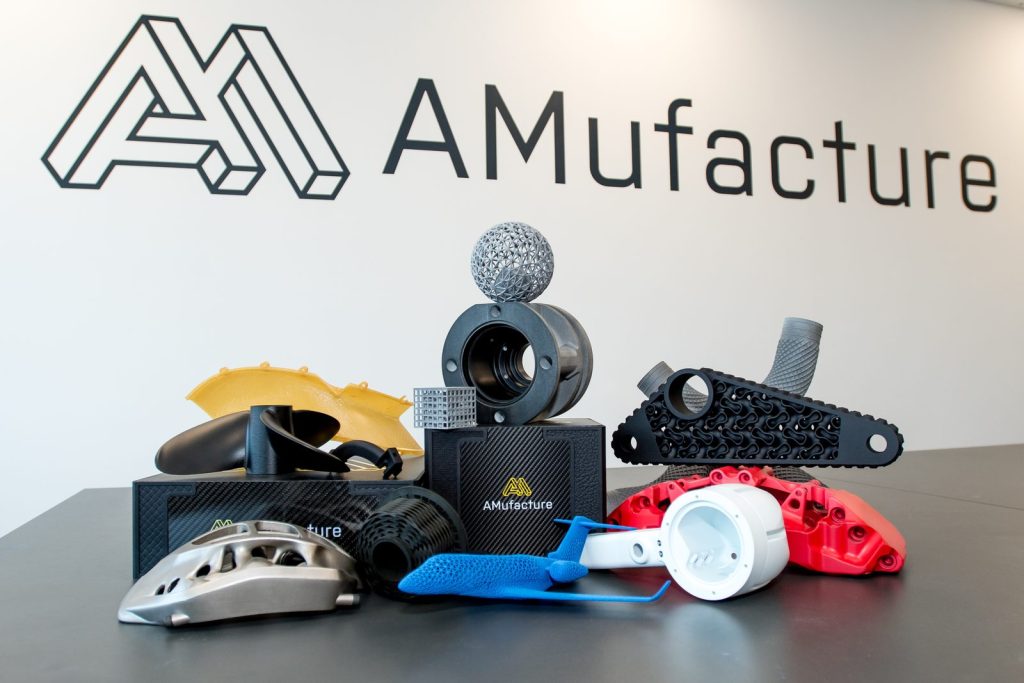 Range of additively manufactured parts, including brake calipers, a propeller and lattice structures, in front of the AMufacture logo