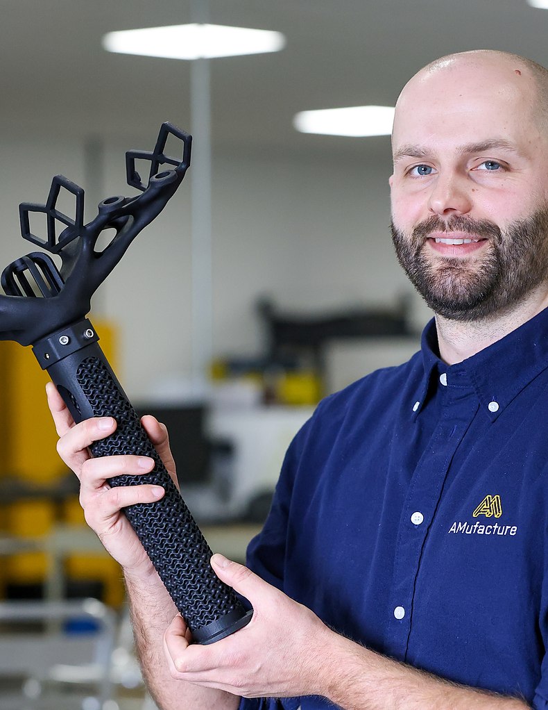 An AMufacture engineer holding a large black 3D printed component on the production floor.