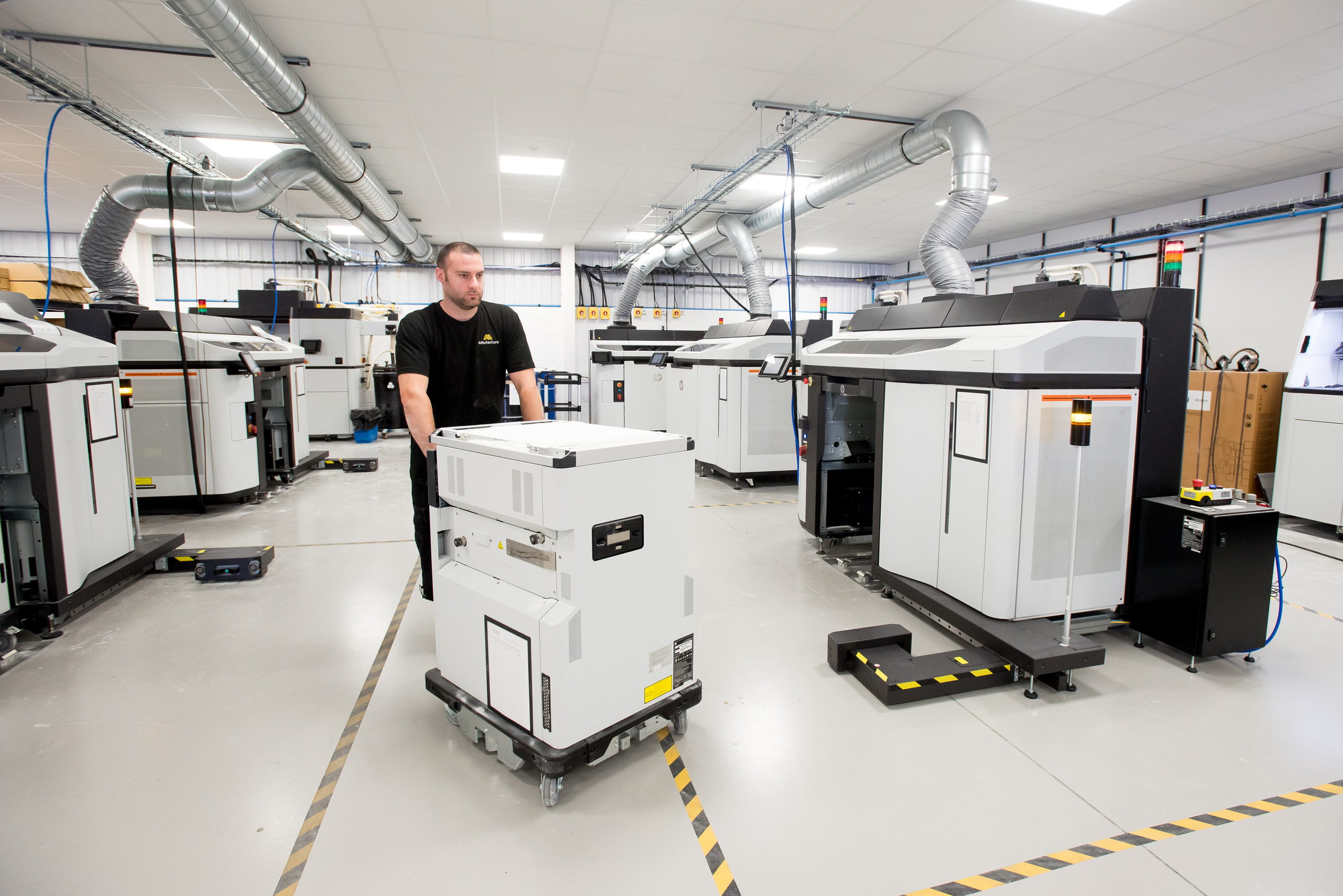 An AMufacture technician wheeling a 3D printer build unit across the production floor, with additive manufacturing machines on either side.