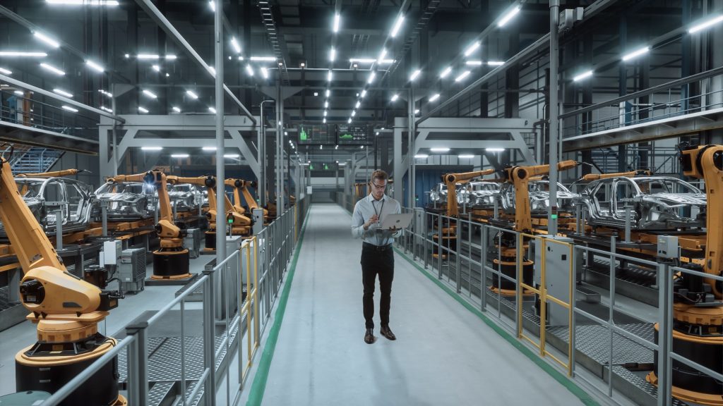 Automotive assembly line with rows of robotic arms building car bodies while an engineer walks the gangway with a laptop