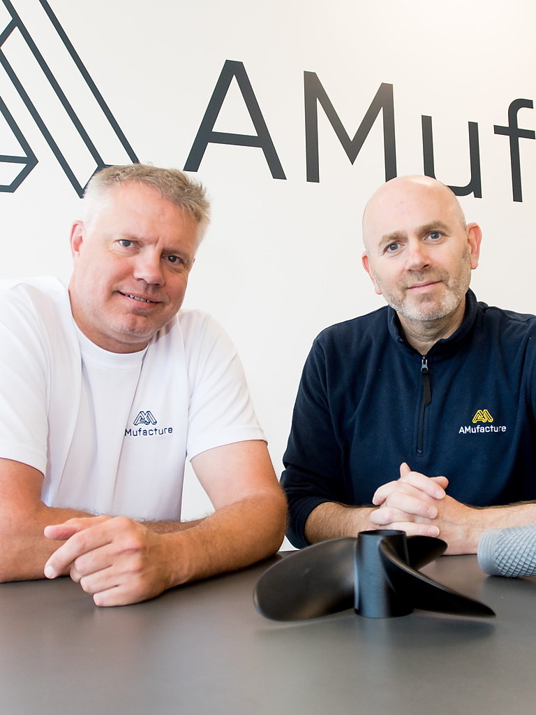 AMufacture co-founders Craig Pyser and Will Howden sat at a table with two 3D printed components in front of them.