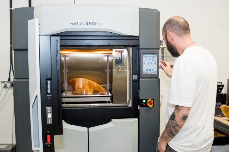 An AMufacture technician at the touchscreen of a Stratasys Fortus 450mc FDM printer, with an orange printed part on the build plate inside the heated chamber