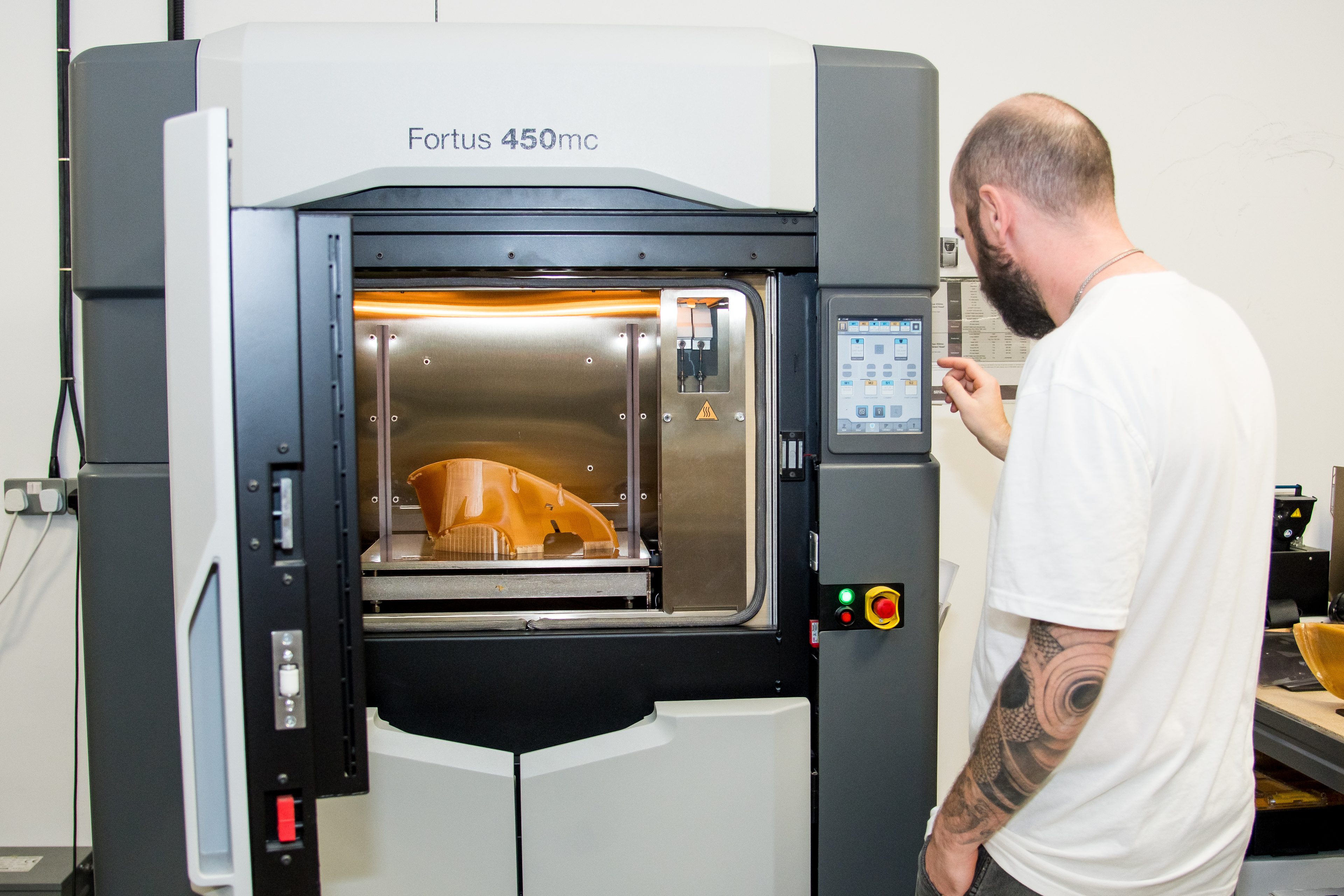 An AMufacture technician at the touchscreen of a Stratasys Fortus 450mc FDM printer, with an orange printed part on the build plate inside the heated chamber